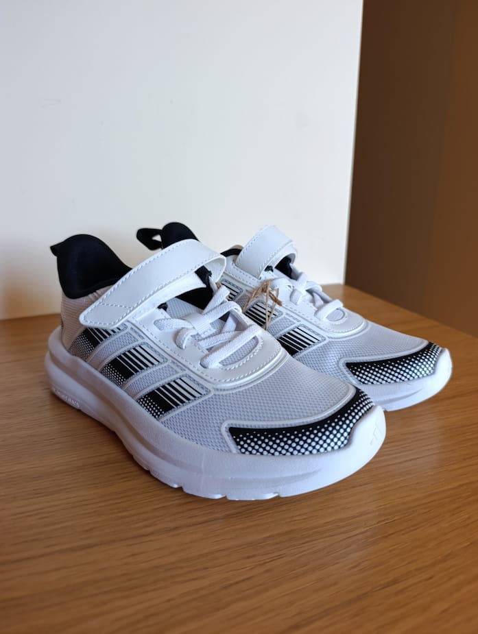 Photo originale d’une paire de chaussures blanches et noires posée sur une surface en bois, avec un cordon d’étiquette accroché aux lacets.