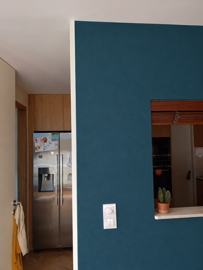 Simulation d’un mur intérieur peint en bleu pétrole, avec interrupteurs, ouverture sur la cuisine et pot de cactus conservés.